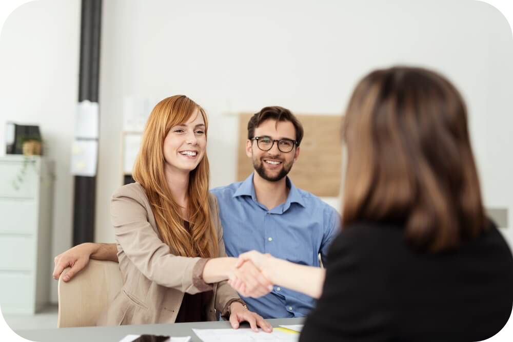 Happy young couple meeting with a broker in her office leaning over the desk to shake hands, view from behind the female agent-modified